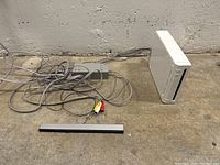 White Nintendo Wii console standing vertically on concrete floor with sensor bar and AV cable laid nearby.