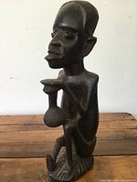 Side profile of dark wooden African figure carving showing kneeling pose and object held in hands.