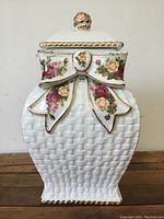 Front view of white ceramic canister with basket weave texture, decorated with pink and yellow roses on a bow design and gold trim on edges and lid knob.