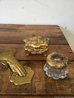 Top angled view showing all three items on a wooden surface: brass hand memo holder, brass jewelry box, and glass lidded container.
