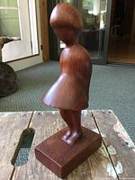 Side view of the solid carved teak wood figure of a girl with a dress and stylized head on wooden base.