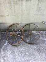 Two antique iron wagon wheels leaning against a wall on a concrete surface, showing overall rust and wear.