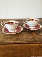 Front view of two Aynsley porcelain tea cups on their matching saucers, showing gold detailing and burgundy bands.