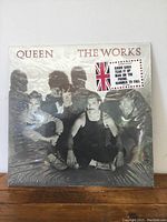 Front cover of the Queen 'The Works' vinyl LP showing the band members with a Union Jack flag sticker listing hit songs.