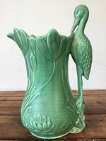 Full view of green SYLVAC pottery jug with a standing crane bird as handle, floral embossed patterns around the body, on a wooden surface.