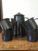 Tall black speckled enamelware coffee pot with lid surrounded by four matching mugs, displaying the complete set from a frontal angle.
