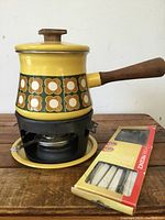 Vintage yellow enamel fondue pot with floral pattern, wooden handle, black metal burner base with fuel container, and yellow drip plate, with box of fondue forks beside it.