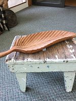 Side view of wooden leaf-shaped dish showing length, carved detail, and natural wood grain on a small rustic bench.