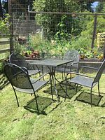 Photo showing square wrought iron patio table with four matching chairs outside on grass near garden.