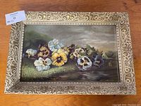 Front view of the antique pansy floral oil painting in its decorative frame on a wooden surface.