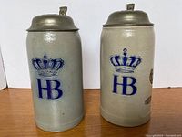 Front view of two beige-gray ceramic beer steins with blue HB logos crowned in blue, showing pewter lids and part of the handles.