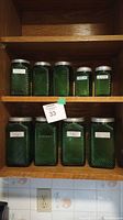 Eight vintage green glass canisters with swirl and textured pattern design, arranged on wooden shelves. Labeled with contents including sugar, flour, cereal, coffee. Metal lids on all.