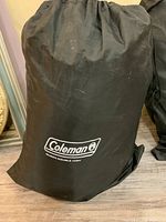 Black storage bag with Coleman logo and text 'QUEEN-DOUBLE HIGH' printed in white, standing upright on a floor surface.