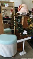 Wide view showing wicker hamper with padded seat, wood plant stand with wreath, pink felt hat on stand, gold-framed mirror, and white jewelry hanger