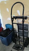 Photo showing black metal hand truck dolly with ball hitch, blue bin with moving blankets, and foldable step ladder against garage wall.