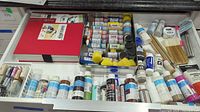 Overhead image showing numerous acrylic paint bottles, brushes, and craft materials laid out on a table.