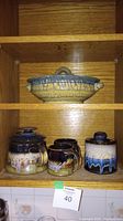 Full view of the lidded tureen on the top shelf and the teapot, two mugs, and sugar bowl on the lower shelf inside a wooden cabinet.