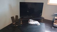 Samsung 32" TV in corner on floor stand with Logitech computer speakers beside it and plastic-wrapped remote on the floor in front.