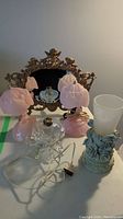 Photo showing all items including two pink glass boudoir lamps, metal cherub mirror with ornate frame, clear glass boudoir lamp base with wiring, and vase with floral resin base.