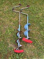 Two manual ice fishing augers lying on grass showing red blade covers and blue shafts with some paint wear and rust on one auger.