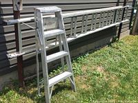 Photo showing the 3-step folding aluminum step ladder standing on grass with siding in the background.