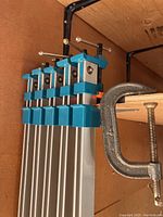 Five large aluminum bar clamps stacked vertically with blue plastic and metal screw handles, clipped to a wooden surface.