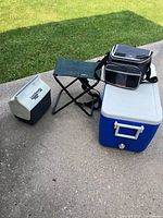 Photo showing three coolers and one folding camp chair arranged on a concrete surface by grass.