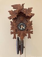 Front view of handcrafted wooden cuckoo clock with carved leaves and bird on top, clock face with Roman numerals, and two pinecone-shaped weights hanging down.