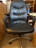 Front view of black leather office chair showing padded seat, back, and armrests, mounted on metal base with wheels.