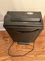 Paper Shredder WMC303X shown on wooden floor, black color, with power cord visible, switch and paper slot on top.