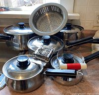 Front view of Lagostina stainless steel pots with lids, steamer insert, egg slicer, pastry blender, and some pot holders and oven mitts on countertop.
