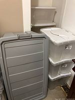 Large gray plastic tote lid leaning against stacked storage totes with white lids.