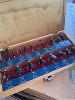 View showing the full set of 20 router bits arranged in the wooden box with blue labels describing each bit profile.