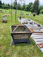 Fire pit with mesh sides and raised base shown outdoors on grass surface with various small tools and items nearby.