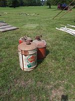 Photo showing three vintage metal gas cans arranged closely on grass with visible rust and branding on cans.
