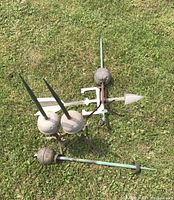 Set of two green lightning rods with concrete bases and metal tripods, smaller metal rod with spherical base, and a weather vane lying on grass.
