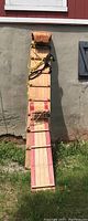 Full length side view of the wooden toboggan showing natural wood and red painted accents along the slats, leaning against a wall outside.
