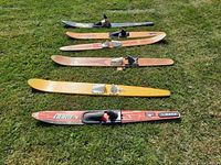 Photo showing six single vintage water skis laid out on grass, various brands visible, including OBrien in red and others in wood and yellow finishes.