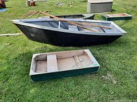 Side view of large black and gray wooden row boat with two oars resting inside, accompanied by a smaller green and white wooden dinghy on grass.