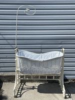 Full view of the antique wrought iron bassinet showing the white quilted liner and vertical support arm against a gray exterior wall.
