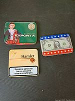 Three cigarette tins shown from top: Macdonald Exporta with woman in Scottish dress, Hamlet miniatures cream tin with health warning label, and a US dollar bill themed tin with stars.