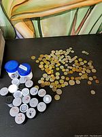 Wide view of assortment of coins spread out on dark surface with several plastic coin containers on the left side.