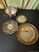 Photo showing the metal pot with two side handles alongside three ornate decorative metal plates of varying sizes with detailed cut-out and engraved designs.
