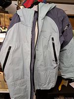 Front view of a two-tone light blue and dark gray outdoor jacket with multiple zippered pockets and a hood.