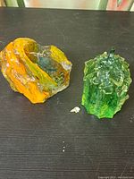 Two art glass sculptures displayed together on black surface - one orange and one green with leaf details.