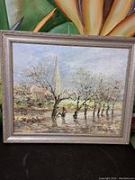 Full view of the oil painting showing the church with tall steeple and trees in front, framed in a silver-colored decorative frame.