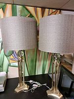 Pair of large metallic gold-toned lamps with fabric drum shades sitting on a table against a colorful backdrop.