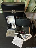 View of the Polaroid 250 camera, manuals, cold clip accessory in box, carrying case, and film pack displayed on a black surface.