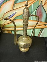 Front view of solid brass coffee decanter showing long curved spout and detailed engraved patterns on body and handle.