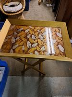 Full shot of the vintage folding tray table showing its wooden frame, glass top, and leaf-patterned surface beneath the glass.